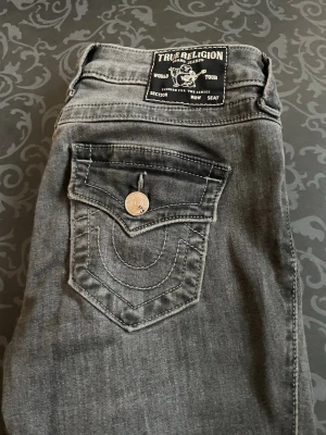 Grå True Religion jeans med fickdetaljer - Snygga grå jeans från True Religion med ikoniska dekorativa fickor med knapp. Klassisk femficksmodell och silverfärgade knappar. Modellen heter BECCA och materialet är mjukt denim. 