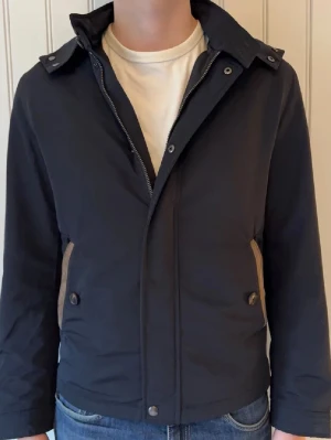 Marinblå vindjacka från Massimo Dutti - Snygg marinblå vindjacka från Massimo Dutti med huva och dragkedja framtill. Jackan har quiltade detaljer och tryckknappar vid kragen. Tillverkad i ett lätt, vindtätt material som passar perfekt för blåsiga dagar. Passar till folk som har S i storlek också