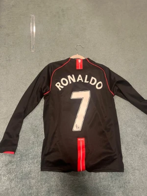 Manchester United Ronaldo tröja svart - Säljer en svart långärmad Manchester United matchtröja från Nike med Ronaldo och nummer 7 på ryggen. Tröjan har röda detaljer, AIG som sponsor på bröstet och klubbmärke på vänster sida. Perfekt för fotbollsfans!
