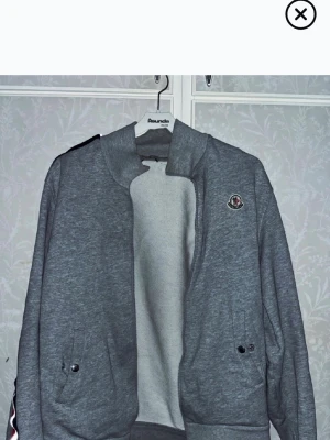 Grå Moncler hoodie med dragkedja - Snygg grå hoodie från Moncler med dragkedja framtill och två fickor med knappstängning. Klassisk Moncler-logga på bröstet och ribbade muddar. Perfekt för en avslappnad och trendig look. Tillverkad i mjukt sweatshirtmaterial.