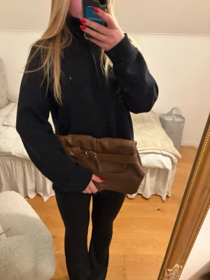 Brun kuvertväska med krokopräglat mönster - Snygg brun kuvertväska i fejkläder med krokopräglat mönster och guldfärgat spänne framtill. Väskan har en vikbar design och ett brett band som detalj. Perfekt accessoar för att lyfta din outfit.