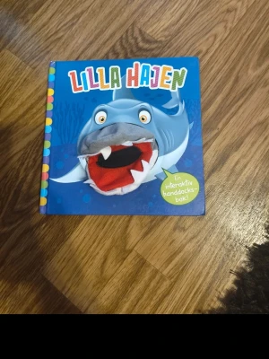 Lilla Hajen interaktiv handdockebok - En färgglad barnbok med titeln 'Lilla Hajen' där hajen har en inbyggd handdocka i tyg. Boken har blått omslag med en stor haj och färgglada bokstäver. Perfekt för högläsning och lek.