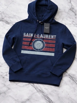 Mörkblå Saint Laurent hoodie - Snygg mörkblå hoodie från Saint Laurent med tryck framtill i rött, vitt och grått. Klassisk huva. Materialet känns mjukt och skönt, perfekt för en avslappnad stil. Saint Laurent-logga och mönster på bröstet.