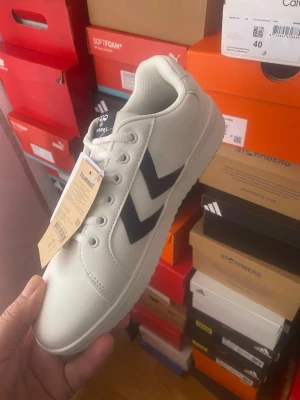 Vita Hummel sneakers med svarta detaljer - Snygga vita sneakers från Hummel med klassiska svarta pilar på sidan. Skorna har lågt skaft, vita snören och en vit sula. Tillverkade i syntetmaterial med textildetaljer och en stilren design som passar till det mesta.