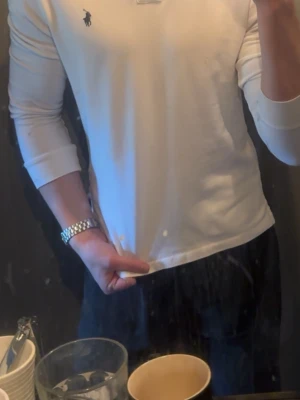 Vit långärmad henley från Ralph Lauren - Säljer en stilren vit långärmad henley från Ralph Lauren med klassisk krage och den ikoniska loggan broderad på bröstet. Skjortan är i mjuk bomull och har en slim passform som sitter snyggt på kroppen. Perfekt för dig som gillar en clean och tidlös look.