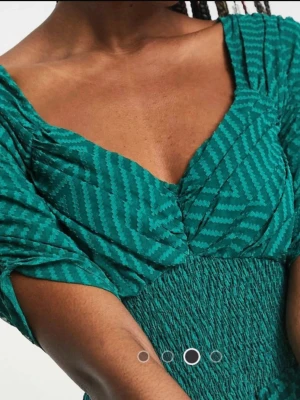 Grön mönstrad midiklänning från Asos - Snygg grön midiklänning med randigt mönster och smock i midjan. Klänningen har v-ringad hals, korta ärmar och ett luftigt, svepande tyg som ger en härlig siluett. Perfekt för dig som vill ha en unik och trendig look.