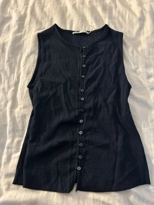 Mörkblå ärmlös linnetopp med knappar - Stilren mörkblå ärmlös linnetopp med knappar hela vägen framtill. Toppen har en lätt strukturerad yta och rund halsringning. Perfekt att matcha med jeans eller kjol för en enkel och snygg look.