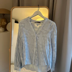 Ljusblå blus med blommönster - Snygg ljusblå blus med diskret blommönster och v-ringning. Blusen har knappar framtill och långa ärmar med lätt puff och resår vid ärmslut. Tillverkad i ett tunt, transparent tyg som ger en luftig känsla. Perfekt att styla med jeans eller kjol. Passar mig som har S/M. Passar även större