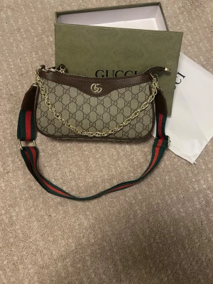 Gucci GG Supreme axelremsväska - Säljer en snygg Gucci GG Supreme handväska i beige och brunt med klassiskt GG-mönster. Väskan har guldfärgad kedjedetalj, justerbar axelrem i grönt och rött samt detaljer i brunt läder. Perfekt för dig som gillar lyxiga accessoarer och vill sticka ut.