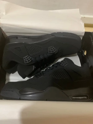 Svarta Air Jordan 4 Retro sneakers - Säljer ett par helsvarta Air Jordan 4 Retro sneakers med ikonisk Jumpman-logga på plösen. Skorna har meshdetaljer, plastpaneler och synlig Air-enhet i sulan. Tillverkade i en mix av läder och syntetmaterial för en clean och sportig look.