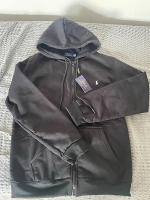  Ralph Lauren hoddie med dragkedja  - Svart hoodie med dragkedja från Polo Ralph Lauren. Klassisk design med huva, fickor framtill och den ikoniska vita broderade loggan på bröstet. Perfekt för en avslappnad och stilren look. Mjuk bomullsblandning och ribbade muddar. Märkt som M men passar som S. Vid mått kom privat 😊