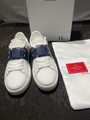 Valentino Garavani vita och blå sneakers - Säljer ett par snygga Valentino Garavani sneakers i vitt med marinblå detalj över snörningen. Skorna har rund tå, platt sula och är tillverkade i skinn. Klassisk design med vita snören och mönstrad yttersula. Perfekt för dig som gillar exklusiva märken.