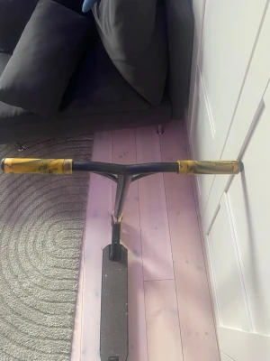 Svart trick sparkcykel med guld detaljer - Säljer en svart trick sparkcykel med guldfärgade handtag och hjul. Styret är rakt och har en robust design, perfekt för tricks och skatepark. Deckytan är greppig och hjulen är små och slitstarka. Passar dig som gillar att åka och göra tricks.