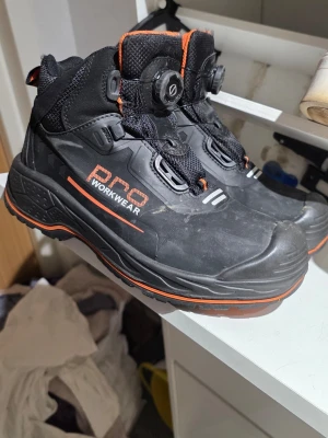 Svarta kängor från Pro Workwear - Säljer ett par svarta kängor från Pro Workwear med orange detaljer och snörning med snabblås. Kängorna har robust sula, förstärkt tå och vadderad insida i orange mesh. Perfekta för dig som vill ha slitstarka och bekväma skor till jobbet.