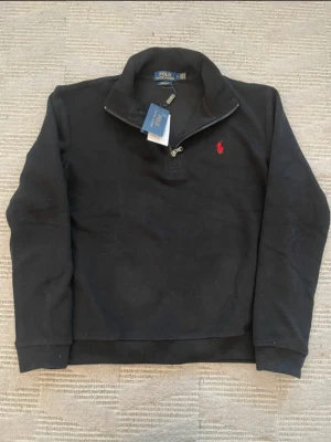 Svart half-zip hoodie från Polo Ralph Lauren - Svart hoodie från Polo Ralph Lauren med half-zip dragkedja och klassisk röd logga broderad på bröstet. Tröjan har hög krage, långa ärmar och ribbade muddar. Tillverkad i mjukt material som känns skönt mot huden. Perfekt för en avslappnad och stilren look.   TVEKA INTE ATT SKICKA BUD 