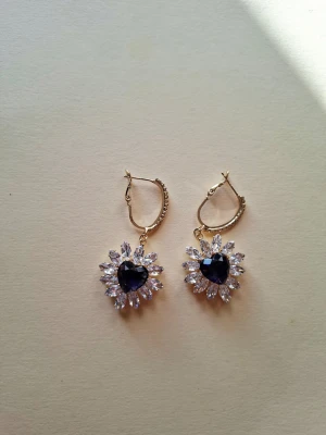 Guldörhängen med hjärtformad sten - Snygga örhängen i guldton med en stor, mörkblå hjärtformad sten i mitten. Runt stenen sitter gnistrande klara stenar i en blomliknande form. Örhängena har klicklås och ger en lyxig känsla med sin kombination av färger och former.