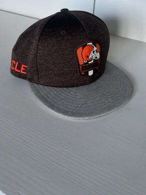 Cleveland Browns SnapBack keps - Snygg snapback keps från New Era med Cleveland Browns-logga framtill. Kepsen är mörkbrun med grått platt brätte och har broderad text i orange på sidan. Tillverkad i ett mjukt och bekvämt material, perfekt för dig som gillar sportig stil.