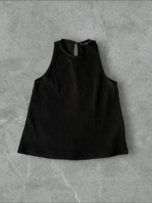 Svart ribbad ärmlös linnetopp - Stilren svart linnetopp med ribbad struktur och hög hals. Toppen är ärmlös och har en enkel, clean design som passar perfekt till många olika outfits. Perfekt för dig som gillar minimalistisk stil.