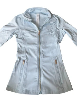 Ljusblå define jacket från Lululemon - Snygg ljusblå define jacket som är exakt likadan som Lululemon med figurnära passform, hög krage och dragkedja framtill. Jackan har två fickor med dragkedja och är tillverkad i ett lätt, stretchigt material som passar perfekt för blåsiga dagar.