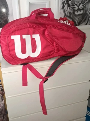 Röd Wilson sportväska - Säljer en röd sportväska från Wilson med stort vitt W-tryck på sidan. Väskan har flera fack med dragkedja och justerbar axelrem. Perfekt för att bära sportutrustning eller använda som gymväska. Tillverkad i slitstark syntet.