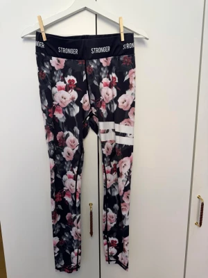 Blommiga tights från Stronger - Säljer ett par snygga träningstights från Stronger i storlek medium. De har ett svart basfärg med rosa och vita blommor samt vita ränder på ena benet. Tightsen har hög midja och är tillverkade i ett stretchigt funktionsmaterial.
