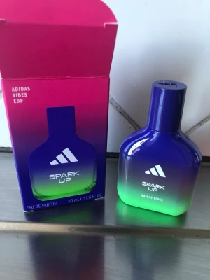 Adidas Spark Up Vibes parfym 50ml - Adidas Spark Up Vibes Eau de Parfum i en snygg flaska med blå och grön färgskiftning. Flaskan har en modern, rundad form och rymmer 50 ml. Kommer i matchande förpackning med rosa och lila toner. Perfekt för dig som gillar fräscha och energiska dofter.
