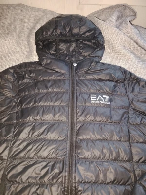 Svart pufferjacka från EA7 Emporio Armani - Snygg svart pufferjacka från EA7 Emporio Armani med huva och dragkedja framtill. Jackan har ett stilrent tryckt logotyp på bröstet och quiltad design. Perfekt för kalla dagar och ger en sportig vibe.