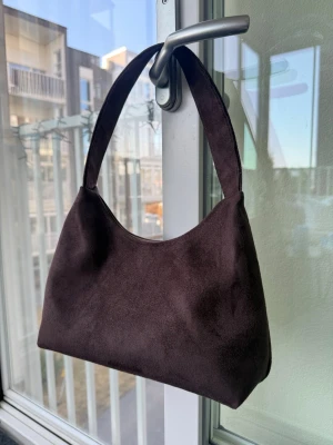 Mörkbrun axelväska i mocka - Stilren mörkbrun axelväska i mjuk konstmocka med enkel, avskalad design och bred axelrem. Väskan har en klassisk rektangulär form och passar perfekt till dig som gillar minimalistisk stil. Storlek ca 26x15cm