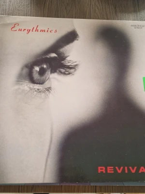 Eurythmics Revival 12" Maxi-singel - Säljer en 12-tums maxi-singel med Eurythmics låt 'Revival'. Omslaget har ett svartvitt foto av ett öga och text i rött. På baksidan finns låtlista och information om mixar och producenter. Perfekt för samlare av vinyl och 80-talsmusik. Inga repor.