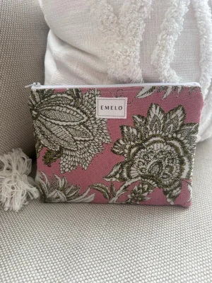 Rosa blommig necessär från Emelo - Snygg necessär från Emelo i rosa tyg med stora blommönster i grönt och vitt. Väskan har en rektangulär form och stängs med vit dragkedja. Perfekt för smink eller småprylar. Insidan är ljus och enkel att hålla ren. Mått ca 20x15 cm