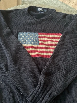 Svart stickad tröja Ralph Lauren - Stickad svart tröja från Polo Ralph Lauren med en stor amerikansk flagga på bröstet. Tröjan har rund halsringning och långa ärmar. Perfekt för dig som gillar klassisk och tidlös stil.