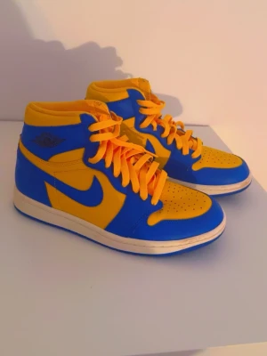 Nike Air Jordan 1 Mid blå/gul - Säljer ett par Nike Air Jordan 1 Mid sneakers i blått och gult. Skorna har klassisk siluett med blå swoosh, gula paneler och matchande gula skosnören. Ovandelen är i läder och yttersulan är vit med blå detaljer. Perfekta för dig som vill sticka ut med färg.