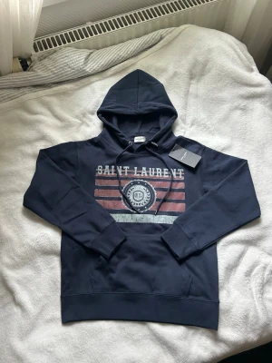 Mörkblå Saint Laurent hoodie - Snygg mörkblå hoodie från Saint Laurent med tryck framtill i vitt och rött som ger en vintagekänsla. Klassisk huva med dragsko och stor magficka. Tillverkad i mjukt bomullsmaterial som är skönt mot huden. Perfekt för en avslappnad och trendig look. Mått finns på bilder!