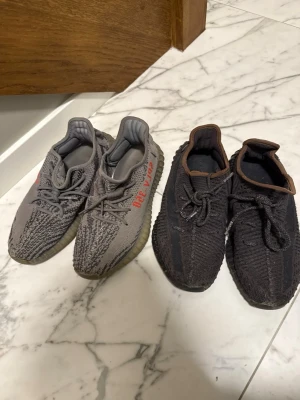 Yeezy Boost 350 V2 sneakers grå & svart - Två par Yeezy Boost 350 V2 sneakers. Ena paret är grått med orange text 'SPLY-350' på sidan och mönstrad ovandel i mesh. Det andra paret är svart med bruna detaljer och samma stickade material. Båda har tjock, räfflad sula och ikonisk siluett.