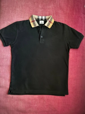 Svart Burberry pikétröja med rutig krage - Klassisk svart pikétröja från Burberry med ikonisk rutig krage i beige, svart, vitt och rött. Tröjan har korta ärmar, knappar framtill och är tillverkad i bomull. Perfekt för en stilren och trendig look. Priset är inte hugget i sten och går att diskutera!