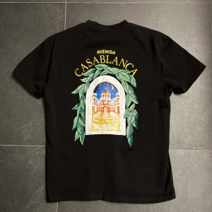 Svart Casablanca Avenida t-shirt - Snygg svart t-shirt från Casablanca med färgglad Avenida Casablanca-print både fram och bak. Trycket visar en byggnad med gröna blad runt, och texten är i gult. Klassisk passform och rund hals. Perfekt för dig som gillar streetwear och unika prints.