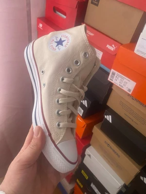 Beige Converse Chuck Taylor All Star - Klassiska beige Converse Chuck Taylor All Star high-top sneakers i canvas med vit tåhätta i gummi, vita snören och sula med röd och blå rand. Ikonisk logga på sidan och metallöljetter för ventilation. Perfekta för en avslappnad och cool stil.