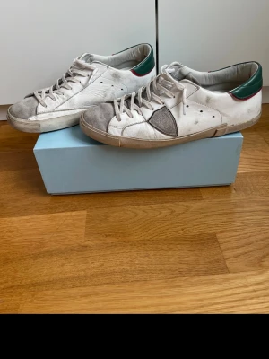 Vita sneakers från Philippe Model - Snygga vita sneakers från Philippe Model med grå mockadetaljer på tån och sidan. Skorna har gröna och röda detaljer vid hälen och klassisk låg profil. Tillverkade i läder och mocka med vita snören och slitstark gummisula. Välandvända men hänger med ett tag till. (Se bilder för skick)