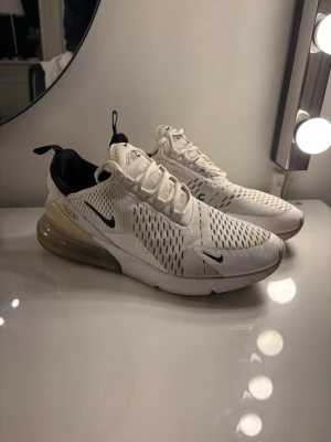 Nike Air Max 270 vita sneakers - Nike Air Max 270 sneakers i vitt med svarta detaljer och stor synlig Air Max-dämpning i hälen. Ovandelen är i mesh för bra andningsförmåga och har en sportig, modern look. Snygga och bekväma sneakers med klassisk Nike-logga på sidan.