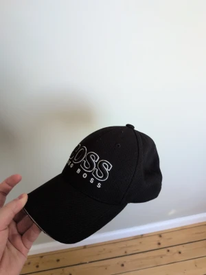 Svart keps från Hugo Boss - Snygg svart keps från Hugo Boss med böjd skärm och broderad logga framtill i vitt. Klassisk modell med justerbar rem bak och ventilerande hål. Tillverkad i ett tåligt tyg som passar perfekt till streetwear och casual outfits.