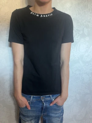 Svart Palm Angels t-shirt - Säljer en svart t-shirt med texten Palm Angels med logga i vitt tryck runt halsringningen. T-shirten har korta ärmar och en klassisk passform. Perfekt för dig som gillar streetwear och vill ha en enkel men snygg look.