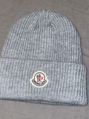 Grå ribbstickad Moncler mössa - Säljer en grå ribbstickad mössa från Moncler med uppvikt kant och klassisk Moncler-logga framtill. Mössan är tillverkad i mjukt, värmande material och har en stilren, enkel design som passar perfekt till kalla dagar.