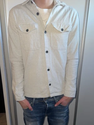Beige overshirt från Jack and Jones  - Stilren beige overshirt med svarta knappar och två bröstfickor med lock. Knappen längst ner är halvt trasig annars inga andra fel med den.