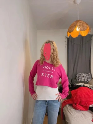 Rosa cropped hoodie från Hollister - Säljer en rosa cropped hoodie från Hollister med vit text på bröstet. Hoodien har långa ärmar och ribbade muddar. Tillverkad i mjukt bomullsmaterial som känns skönt mot huden. Perfekt för en avslappnad och trendig look.