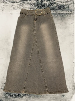  lång jeanskjol med bälte - Säljer en lång jeanskjol med utsvängd passform och coolt bälte med nitar och rund spänne. Kjolen har fickor både fram och bak samt markerade sömmar som ger en snygg vintagekänsla. Perfekt för dig som gillar en avslappnad men trendig stil. Kjolen är lite mer blå än vad den ser ut på bilderna! 
