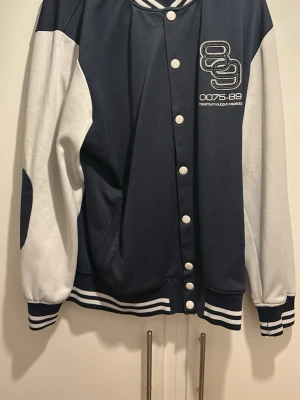 Blåvit bomberjacka med tryck - Snygg blåvit bomberjacka med vita ärmar och mörkblå kropp. Jackan har tryckknappar framtill, randiga muddar och ett broderat tryck på bröstet med siffror och text. Klassisk collegestil som passar perfekt till streetwear.