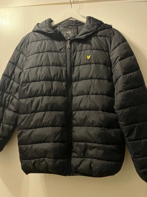 Svart pufferjacka från Lyle & Scott - Säljer en svart pufferjacka från Lyle & Scott med huva och dragkedja framtill. Jackan har det klassiska gula logomärket på bröstet och quiltad design. Perfekt för kalla dagar och riktigt skön att bära. Materialet är syntet och jackan är lätt vadderad.