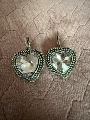 Hjärformade örhängen från Unica - Säljer ett par hjärtformade örhängen från Unica i silverfärgad metall med stora, gnistrande klara stenar i mitten. Örhängena har en detaljerad kant och klassisk klickstängning. Perfekta för dig som gillar statement-smycken med romantisk touch.
