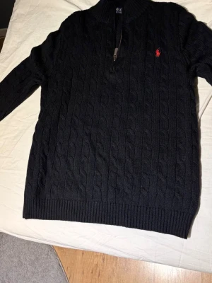 Svart kabelstickad tröja Ralph Lauren - Kabelstickad svart tröja från Polo Ralph Lauren med halv dragkedja och röd broderad logga på bröstet. Tröjan har ribbade muddar vid ärmslut och nederkant samt hög krage. Perfekt för dig som gillar klassisk och stilren design. Obs rep är storleken S men kan även passa XS. Billigare vid snabb affär 