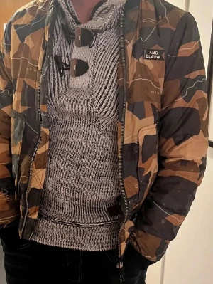 Kamouflage pufferjacka från Scotch & Soda - Säljer en snygg pufferjacka från Scotch & Soda AMS BLAUW med kamouflagemönster i bruna, svarta och beige toner. Jackan har dragkedja, ribbade muddar och en cool streetwear-look. Perfekt för kalla dagar och riktigt skön att bära. Inga defekterna, luvan är avtagbar, skriv vid frågor eller intresse🥰